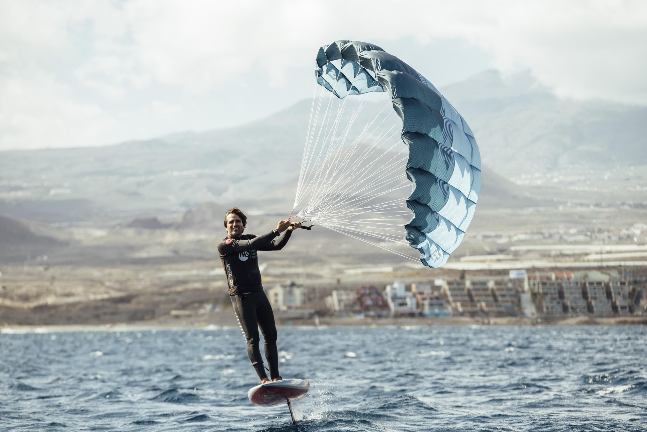 Rider on the Aeryn P1 foiling past a coastal town with mountain backdrop