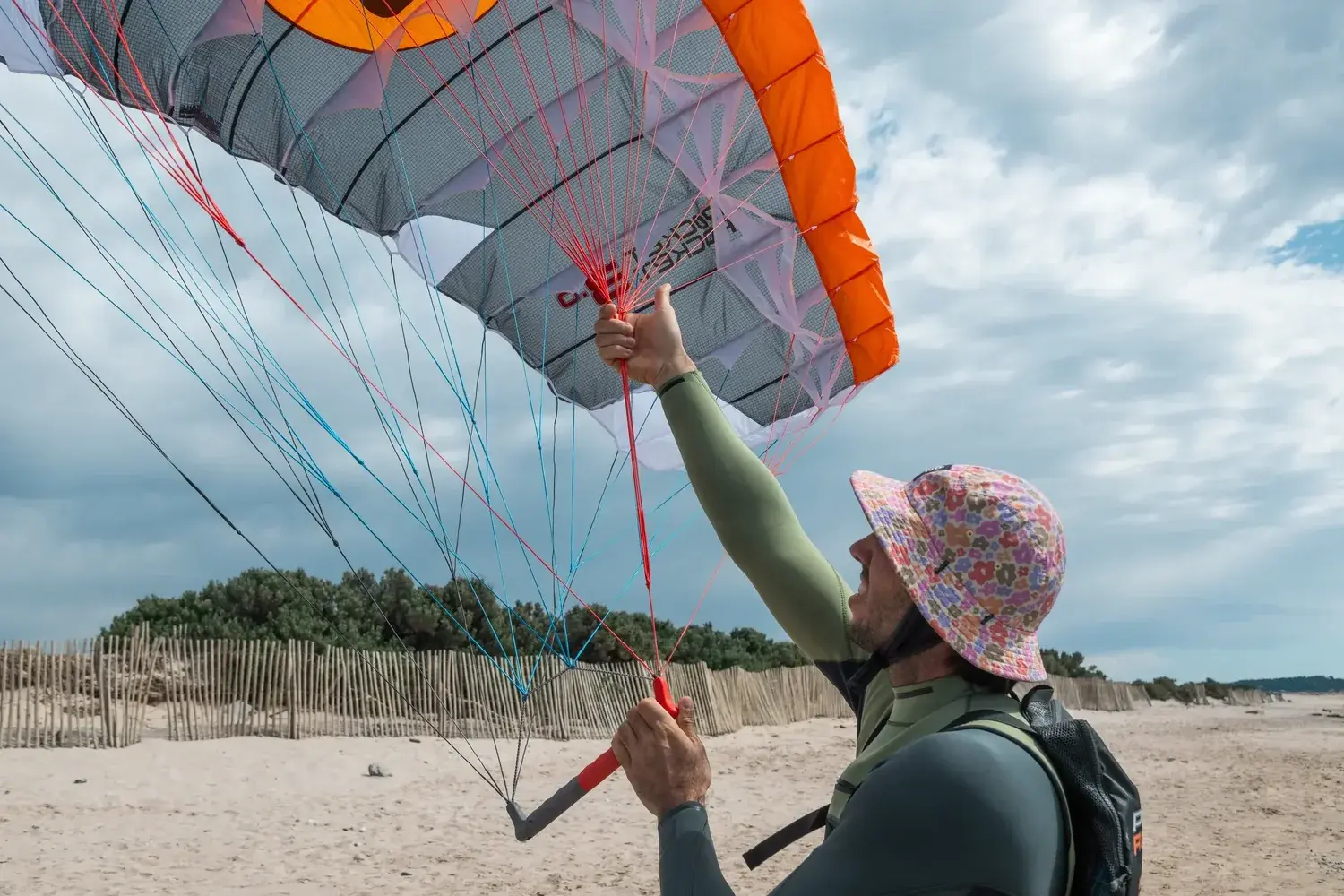 A rider on a sandy beach holding the canopy overhead by its lines, the bridle fanning out in three separate colours, red, blue and black, with the red group gathered into one hand and the control bar hanging from the other