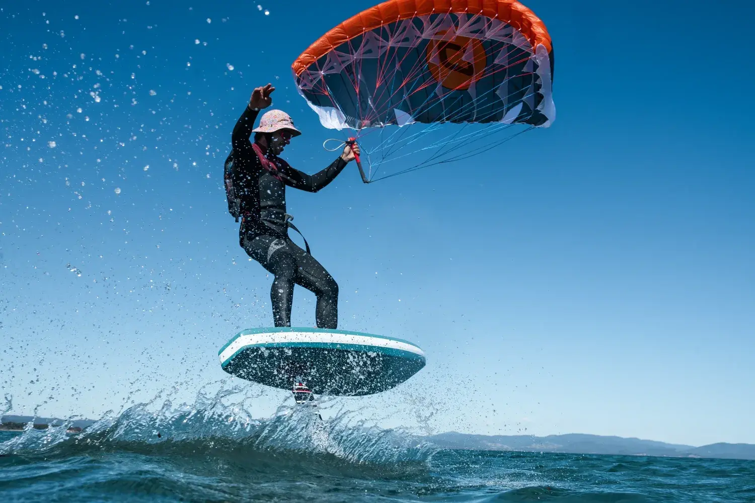 Rider lifting clear of a breaking wave on a foil board, spray thrown across the frame, working the orange and grey canopy overhead with one hand on the bar and the other raised