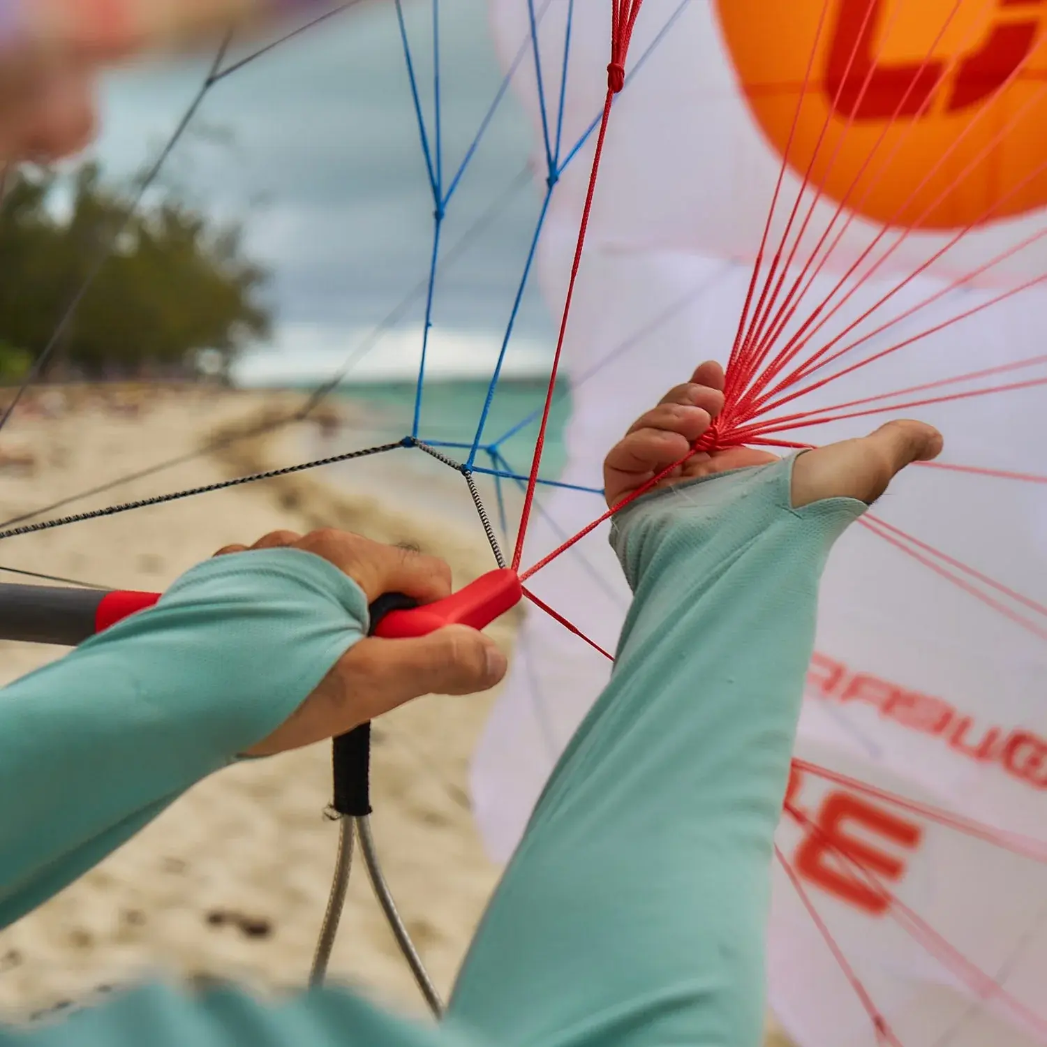 Close-up of a rider's hands on a beach, one gripping the red end of the control bar and the other holding the grouped red centre lines where they fan out, with the white and orange canopy laid out behind