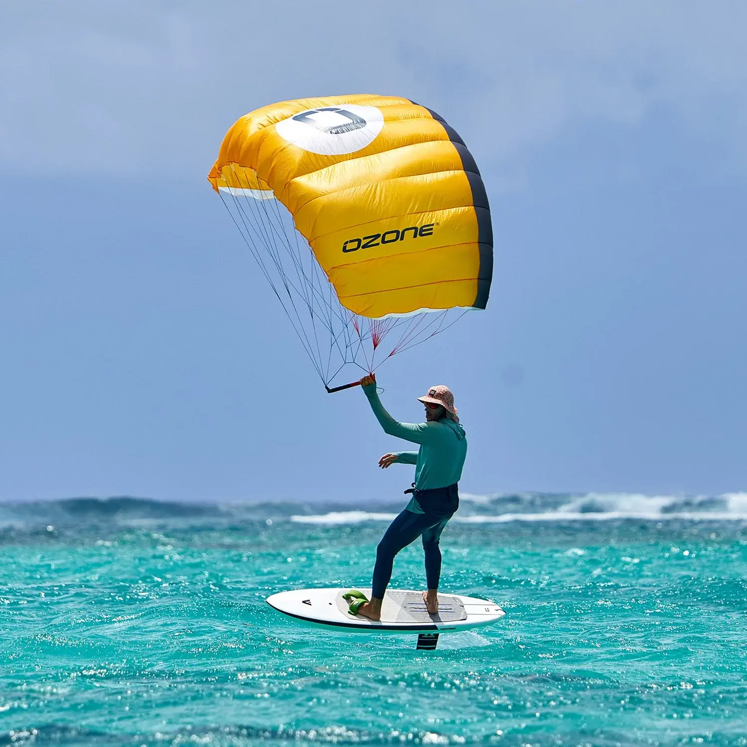 Rider foiling on flat turquoise water under a fully inflated yellow and grey Ozone canopy, holding the bar one-handed, with a breaking reef wave in the distance