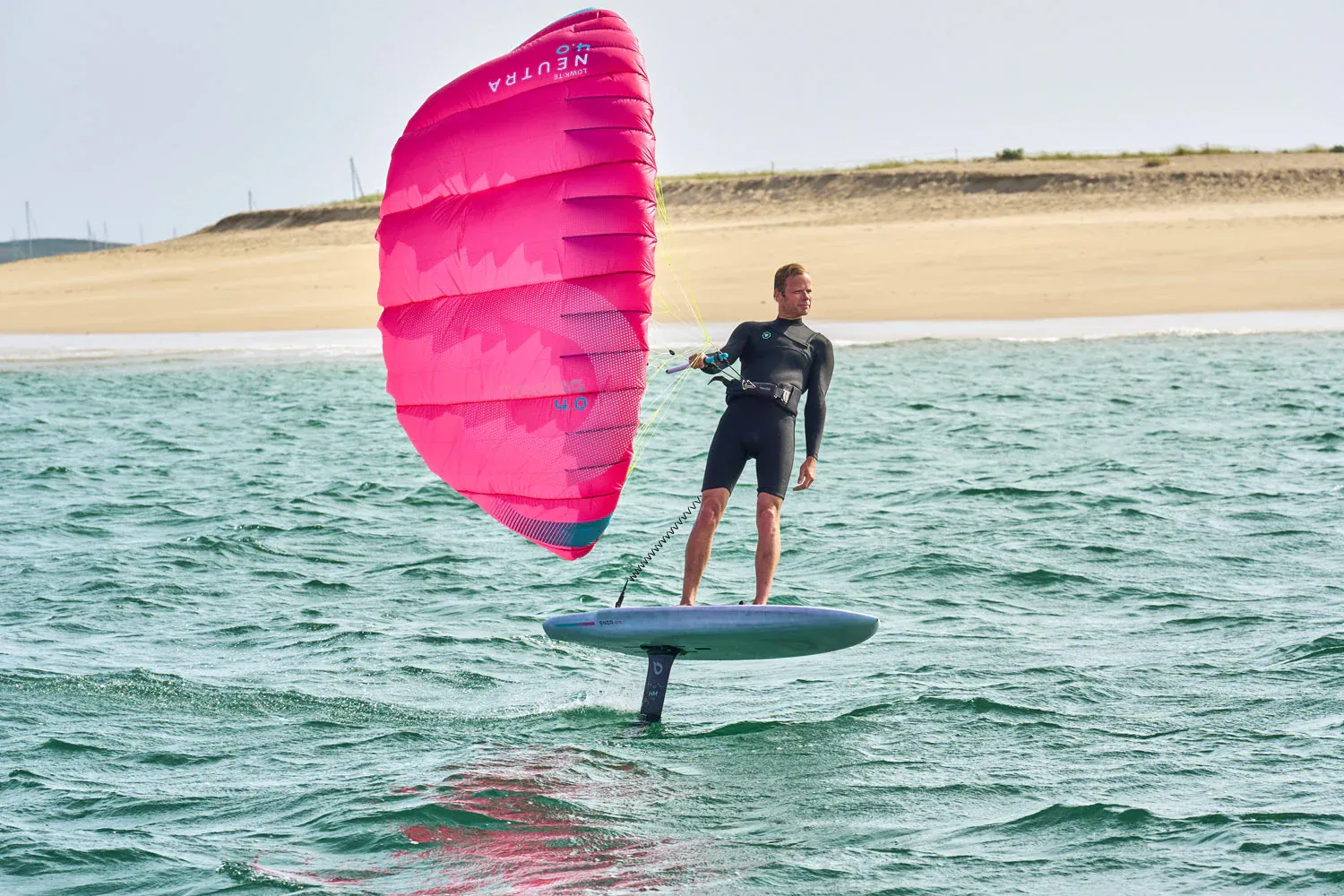 Gong Lowkite rider foiling with pink wing near a sandy beach in light wind conditions