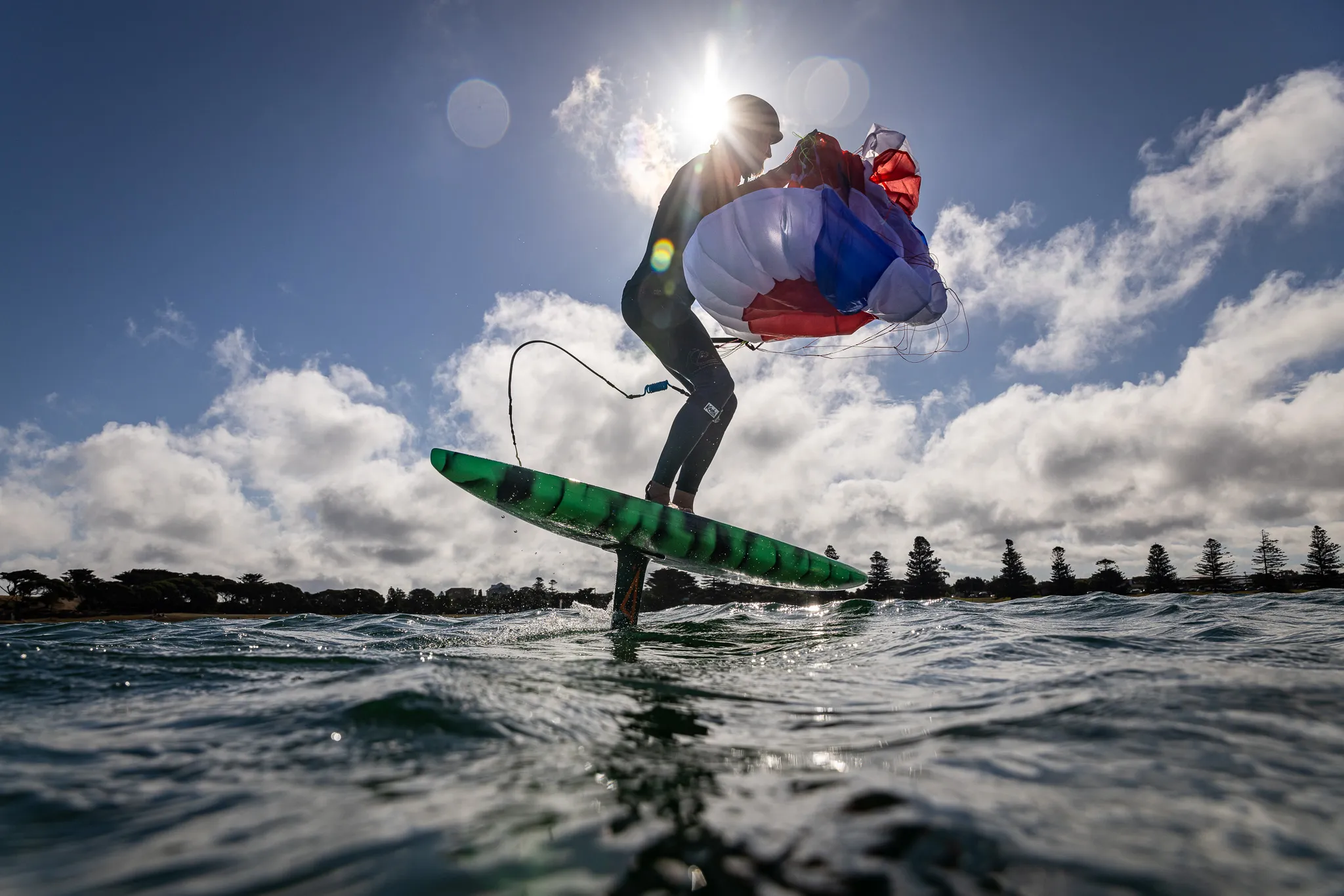 Flow D-Wing rider foiling with the parawing deployed overhead, backlit by the sun creating a dramatic silhouette